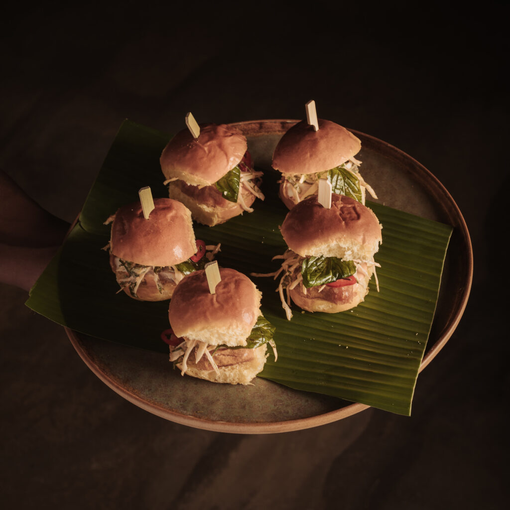 The image displays a beautifully arranged platter of gourmet sliders, served on a large ceramic plate lined with a banana leaf. Each slider is held together with a small wooden skewer and features fresh ingredients like crisp lettuce, slaw, and slices of pickled vegetables, tucked inside soft, golden buns. The presentation is simple yet refined, with an emphasis on fresh, vibrant textures and flavors. The warm lighting highlights the natural tones of the dish, creating a visually appealing composition that emphasizes the care and attention put into the food at Aunty Kim's.