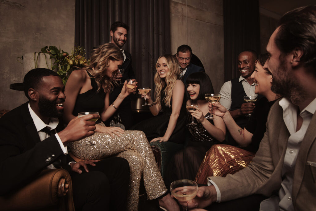 A group of well-dressed friends are gathered closely together, laughing and toasting with glasses of champagne. The group radiates joy and camaraderie, with everyone dressed in chic evening wear, including sequin outfits and tailored suits. The setting is intimate, featuring soft lighting and a moody atmosphere with dark velvet curtains and minimalist decor in the background. The group is seated and standing around a cozy lounge area, capturing a lively celebration filled with conversation and shared moments at this Aunty Kims wedding.