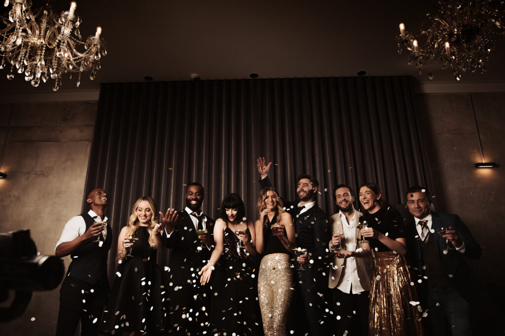 The image shows a lively group of well-dressed people celebrating with enthusiasm at an Aunty Kims wedding. The guests, dressed in glamorous evening wear with shimmering gowns, sleek suits, and tuxedos, are standing under the warm light of chandeliers. Confetti fills the air as they joyfully toss it up, laughing and smiling with drinks in hand. The dark, elegant drapery in the background adds a luxurious touch to the scene, which is brimming with excitement and revelry. The moment captured feels celebratory and carefree, a testament to the festive atmosphere at the venue.