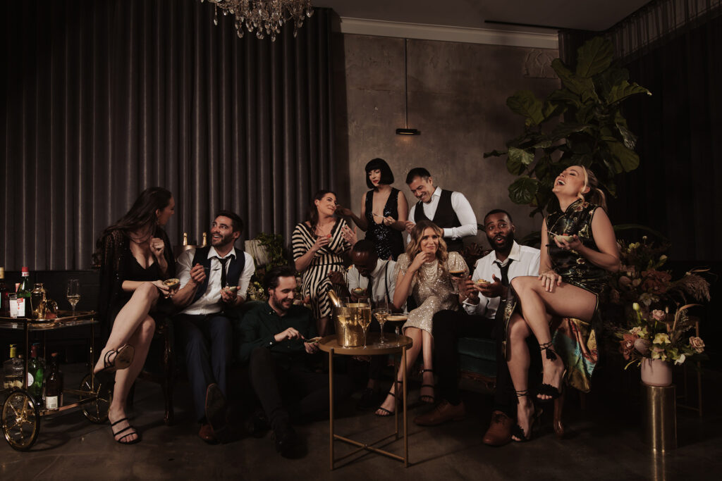 A lively group of friends is gathered in a moody, stylish room at Aunty Kim's. They are dressed in elegant party attire, laughing and chatting while holding drinks and small plates of food. Some are seated on a plush green sofa, while others stand or sit casually on the floor. A small table with champagne glasses sits in the center, while a vintage bar cart with bottles and glasses is nearby. The room is softly lit by a chandelier overhead, and dark curtains and greenery add to the intimate, celebratory atmosphere.