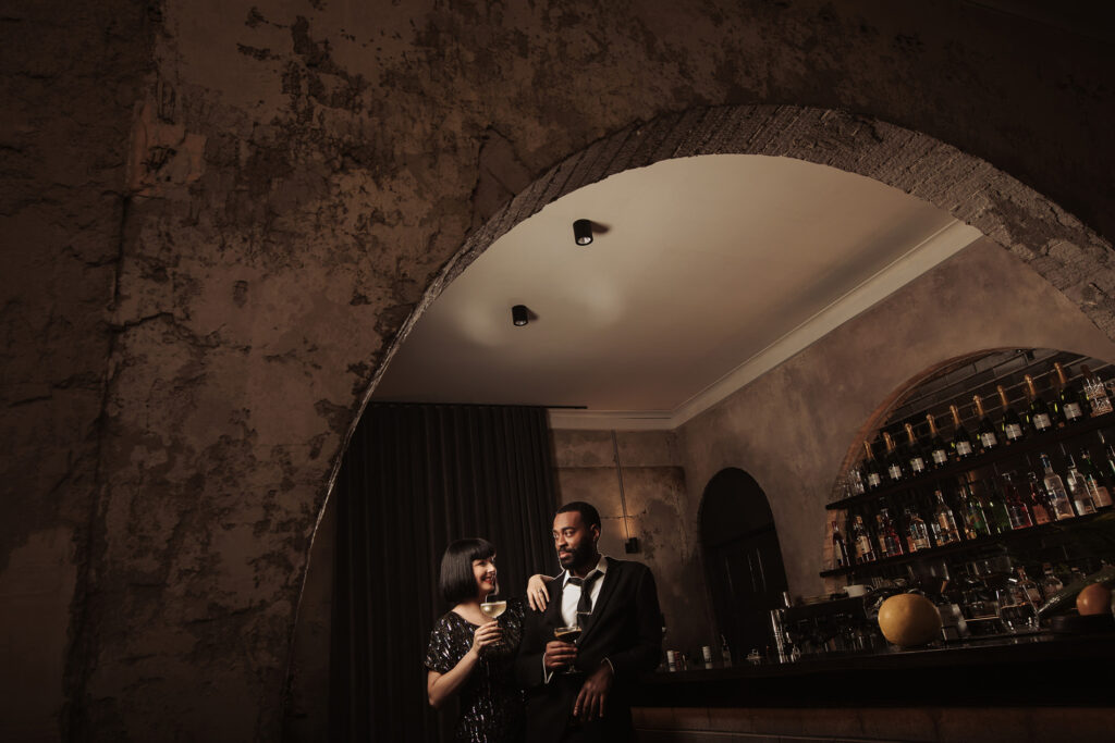 A couple is seen sharing a relaxed moment in an elegant, dimly lit space. The woman is dressed in a sleek black dress with a bob haircut, holding a cocktail and smiling at her companion. The man, wearing a sharp black suit and tie, holds a drink while leaning casually against the bar. The background features an arched, textured wall with exposed details, creating a rustic yet chic atmosphere. Shelves of neatly arranged liquor bottles are visible behind the bar, adding to the setting's refined, vintage vibe. The overall mood is sophisticated and intimate, perfect for a stylish celebration or wedding reception.