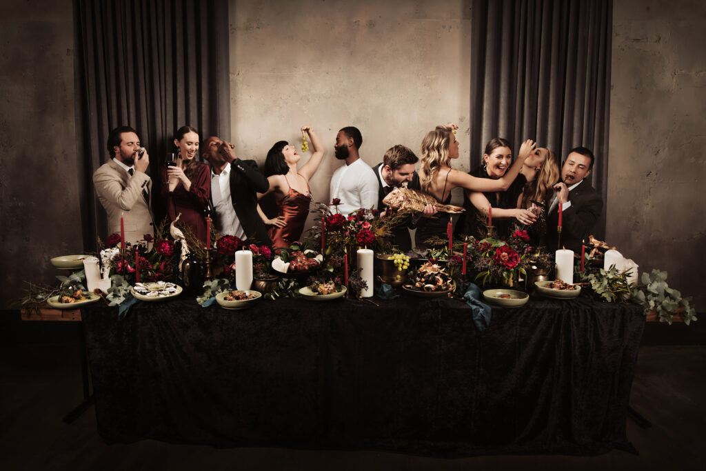 Aunty Kim's wedding venue campaign photoshoot featuring a lavish banquet table adorned with rich floral arrangements and candles, surrounded by a lively group of elegantly dressed guests enjoying food, drinks, and vibrant interactions, against a moody backdrop.
