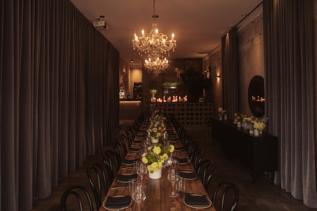 A long wooden dining table is set with black napkins, glassware, and minimalistic white floral centerpieces. Crystal chandeliers hang overhead, casting a warm light over the elegant Aunty Kims wedding space. The walls are lined with soft, dark drapery, and in the background, a bar softly illuminated by candles adds to the intimate ambiance. The scene is refined and atmospheric, designed for a sophisticated dinner or intimate wedding reception.