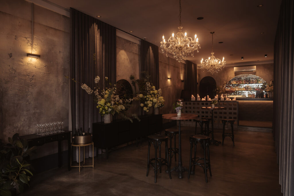 This image shows a dimly lit Aunty Kims wedding and event space with crystal chandeliers, concrete walls, and velvet curtains. Floral arrangements with white and green flowers sit on black sideboards. A bar in the background is softly illuminated, with candles lining the counter. High-top tables and black stools are placed throughout the room, creating a cozy, elegant atmosphere.