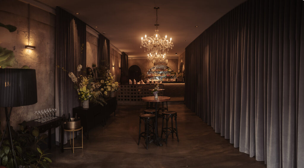 A warm, dimly lit bar at the end of the Aunty Kims wedding space with textured plaster walls and soft, dark drapery on both sides. Crystal chandeliers hang overhead, illuminating the polished concrete floors. Tall floral arrangements add a touch of elegance. A cluster of champagne glasses and bottles sit ready on a side table. The scene is refined and intimate, perfect for a stylish gathering.