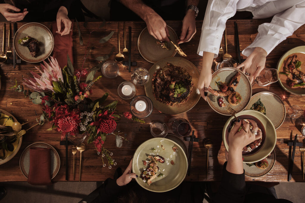 This image appears to showcase a beautifully styled dining table at a wedding reception at Aunty Kim's. The table features a rustic wood surface, decorated with elegant floral arrangements consisting of deep red and pink blooms, accented with lush greenery. The setting is intimate, with guests enjoying gourmet dishes artfully presented on ceramic plates. The atmosphere is warm, with soft candlelight adding to the cozy, yet refined ambiance. The mood suggests a gathering filled with conversation, connection, and the shared experience of exquisite food.