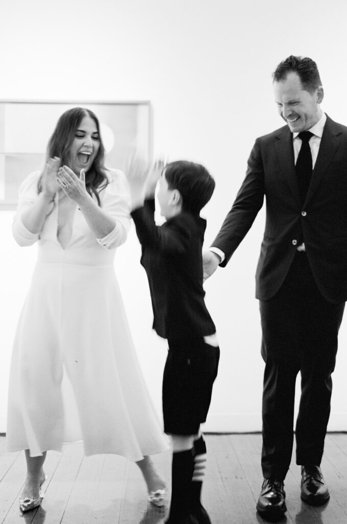 A joyful bride in a flowing white gown from Carte Blanche Bridal is celebrating with a young boy, high-fiving him while laughing. She stands next to a man dressed in a black suit, smiling warmly. The bride’s minimalist dress features a deep V-neckline and subtle detailing, while her jewelled heels add a touch of glamour. The trio shares a lively and playful moment, captured in motion, creating a candid and light-hearted atmosphere.