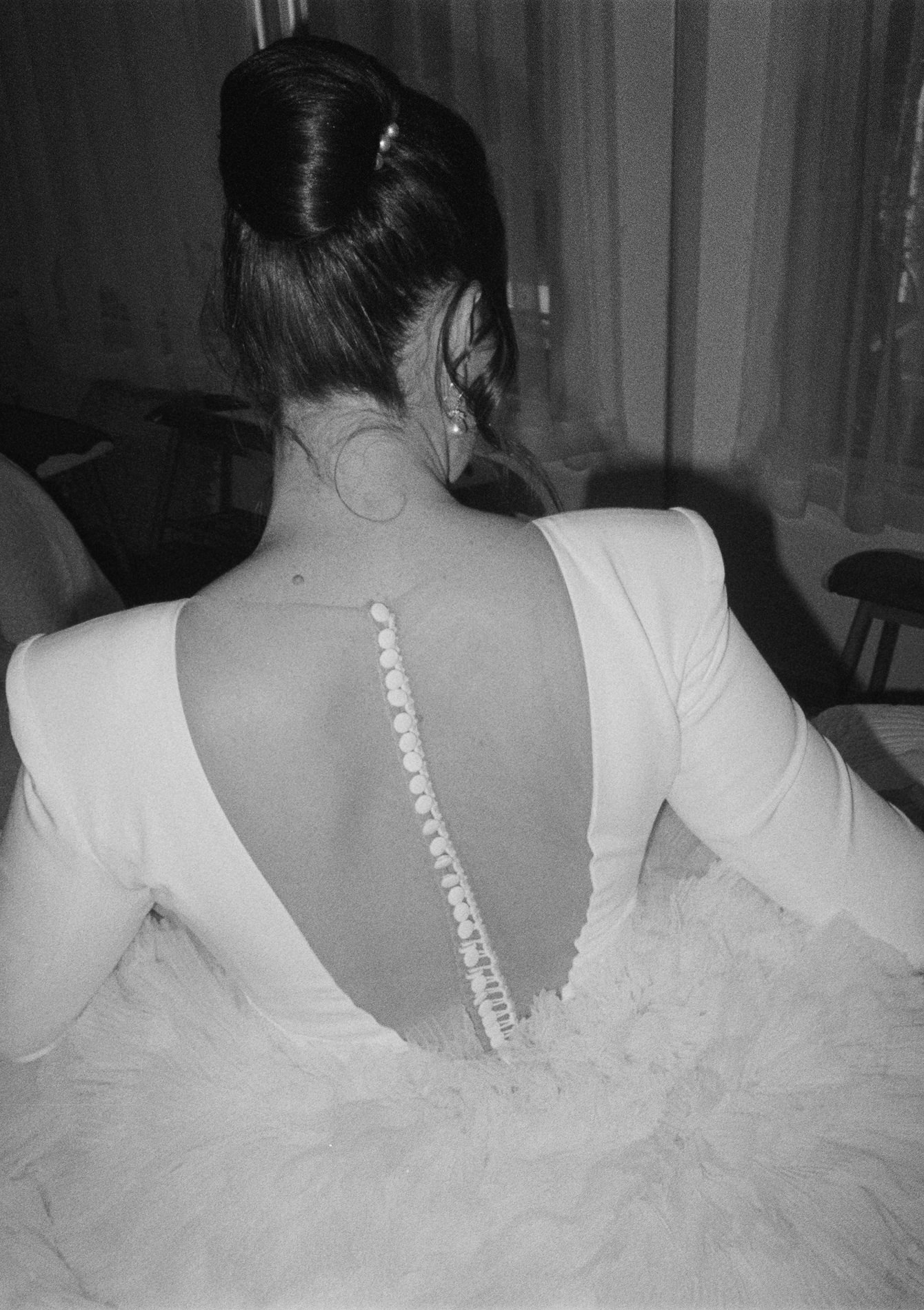 Black and white 35mm film photograph of a bride from behind, showcasing a classic updo with a pearl hairpin and a buttoned back detail on her wedding dress. The image captures the elegant simplicity of the bridal style, emphasizing the texture of the fabric and the timeless quality of film photography.