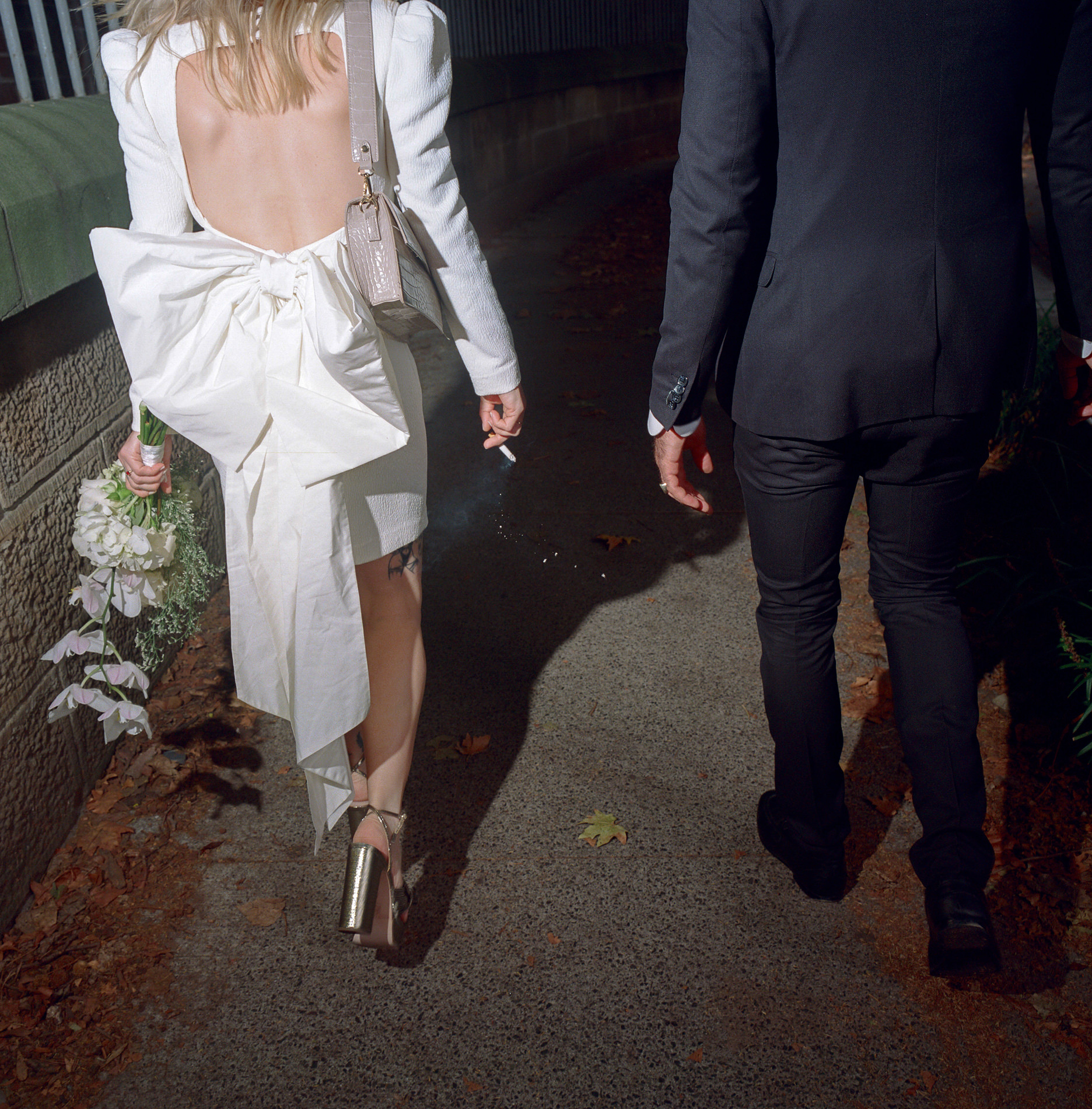 Medium format analog film photograph of a bride and groom walking down a dimly lit path in Sydney. The bride, wearing a chic open-back dress with a large bow detail, holds a bouquet in one hand and a cigarette in the other. The groom, dressed in a tailored suit, walks beside her. The image captures the candid, modern vibe of the couple's wedding day, with the soft evening light casting long shadows on the path.