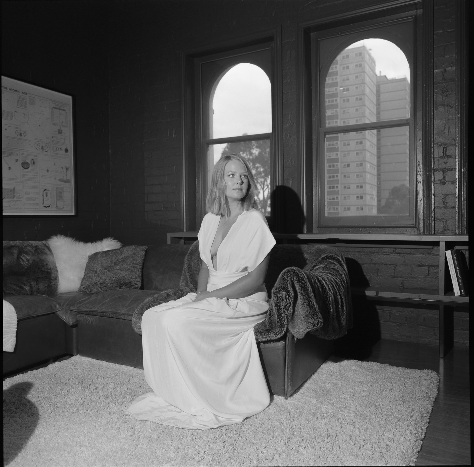 A medium format film photograph taken in Melbourne, capturing a serene moment of a bride seated on a cozy, fur-draped couch in a dimly lit room with brick walls. The bride, wearing a simple, elegant white gown with a deep V-neckline, gazes thoughtfully out the window, where a view of urban high-rise buildings is visible. The image blends the warmth of the interior with the coolness of the cityscape outside, creating a juxtaposition of intimate and urban elements.
