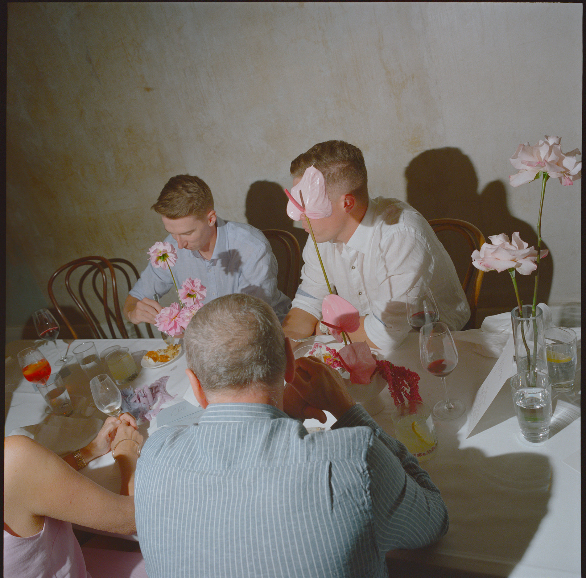 Medium format analog film photograph of wedding guests seated around a table, adorned with vibrant pink flowers. The casual, candid moment captures the guests mid-conversation and dining, with the textured wall in the background adding a rustic charm to the intimate setting.