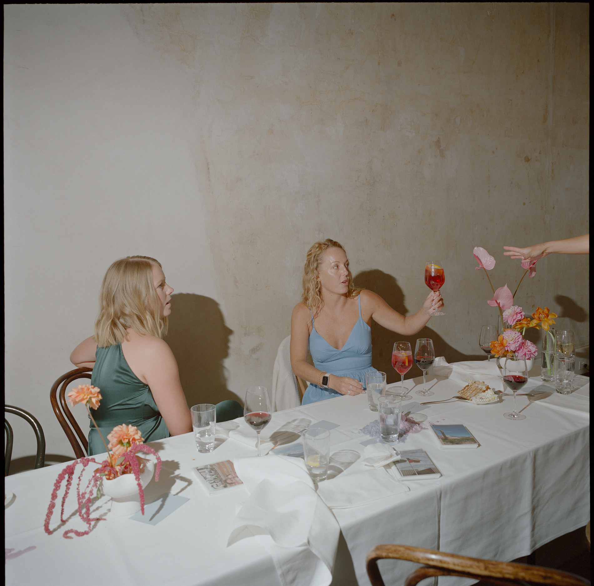 A medium format film photograph taken at a wedding, showing two women seated at a table in a rustic, minimalist setting. One woman, dressed in a light blue dress, raises a glass of red-colored drink as if making a toast, while the other, in a green dress, listens attentively. The table is adorned with a simple floral arrangement, various glasses of wine, and a few scattered items, capturing a candid, relaxed moment of conversation and celebration.
