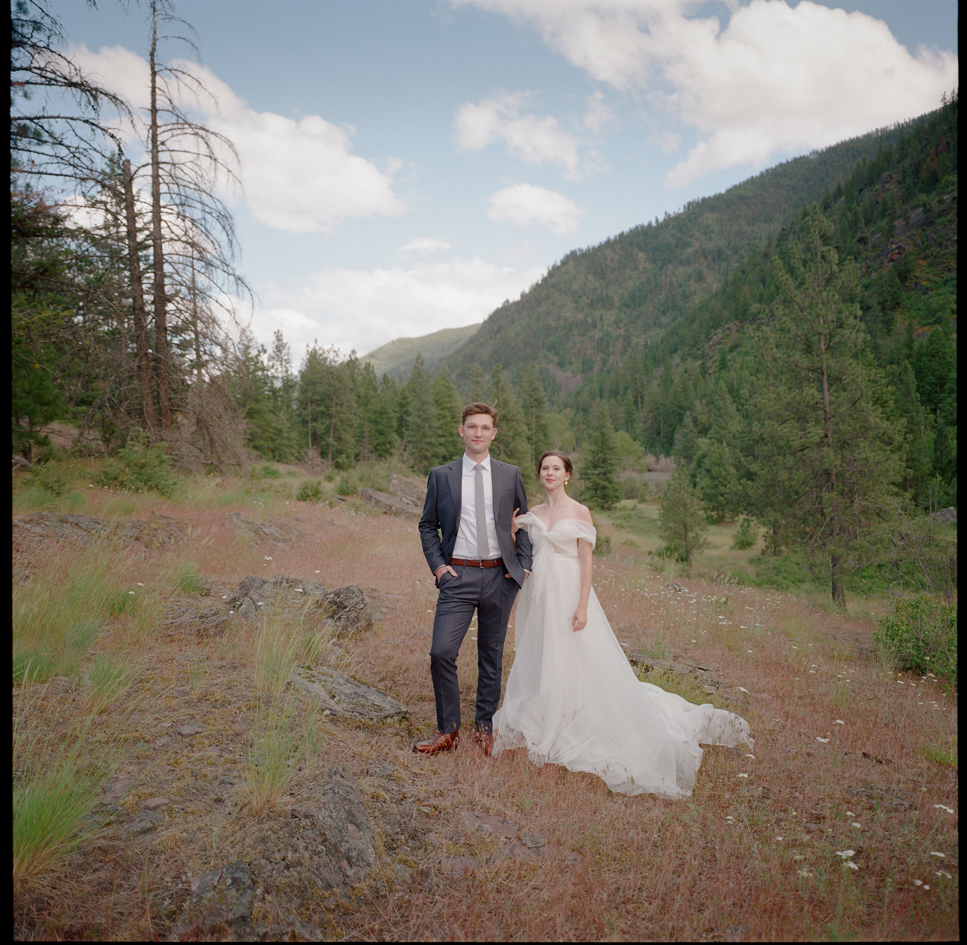 A medium format analog film photograph of a bride and groom standing in a picturesque outdoor setting surrounded by mountains and pine trees. The bride wears an off-the-shoulder wedding dress while the groom is dressed in a tailored suit. The serene landscape enhances the timeless and natural feel of the image, capturing the couple's elegance against the backdrop of nature.