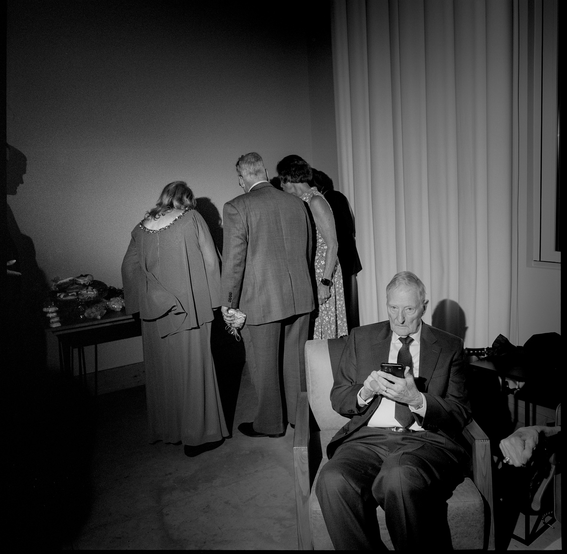 A black-and-white medium format film photograph taken at a wedding in Montana, USA. The image captures an older man sitting alone on a chair, engrossed in his phone, while a small group of guests, dressed in formal attire, gathers in the background around a table. The contrast between the solitary figure and the group highlights a candid moment of quiet amidst the social event.