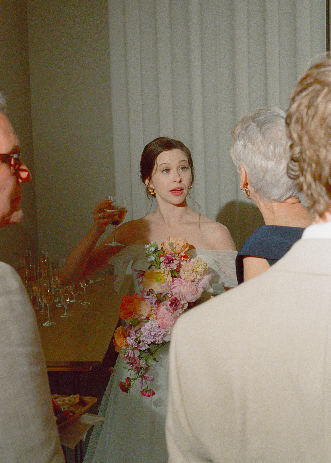 A candid moment captured at a wedding in Montana, USA, featuring a bride in an off-shoulder gown with a vibrant, cascading bouquet of colorful flowers. The bride is holding a glass of wine, engaging in conversation with guests who surround her, all bathed in soft, warm indoor lighting.