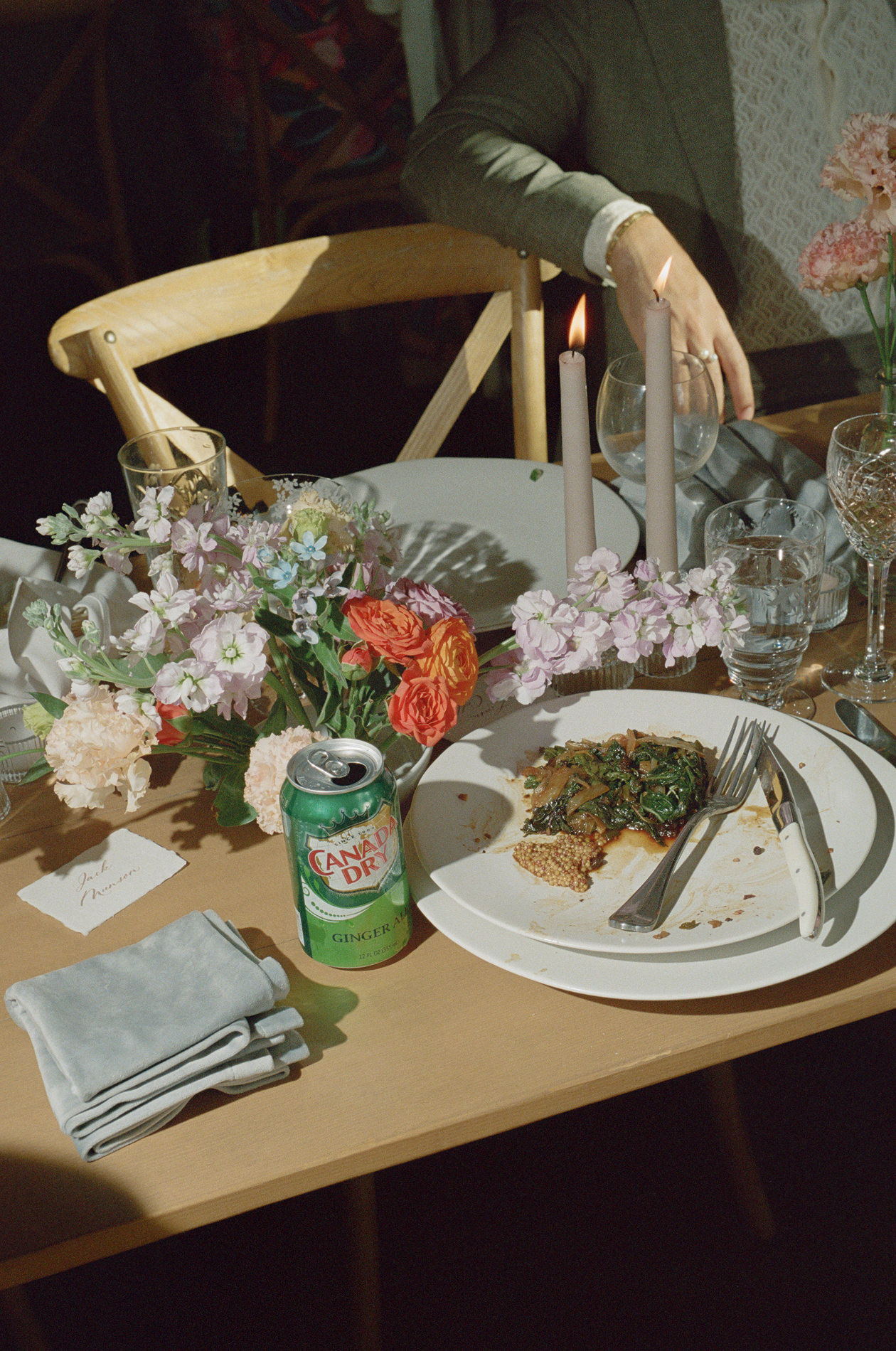 A 35mm film photograph capturing a partially eaten meal at a wedding, with a plate of food and a can of Canada Dry ginger ale on the table. The table is adorned with a colorful floral arrangement, lit candles, and glassware, creating a charming yet casual setting. A folded napkin and a name card reading 'Jack Morrison' are placed beside the plate, hinting at the personalized details of the event.