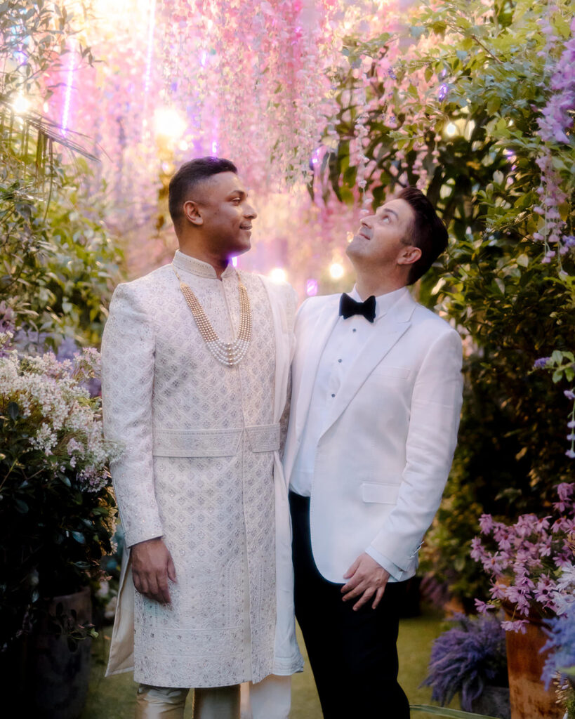 Damian and John at their wedding at one of the most luxury wedding venues Sydney has on offer, The Grounds of Alexandria. Damian and John stand together underneath a brightly coloured pink and purple floral installation.