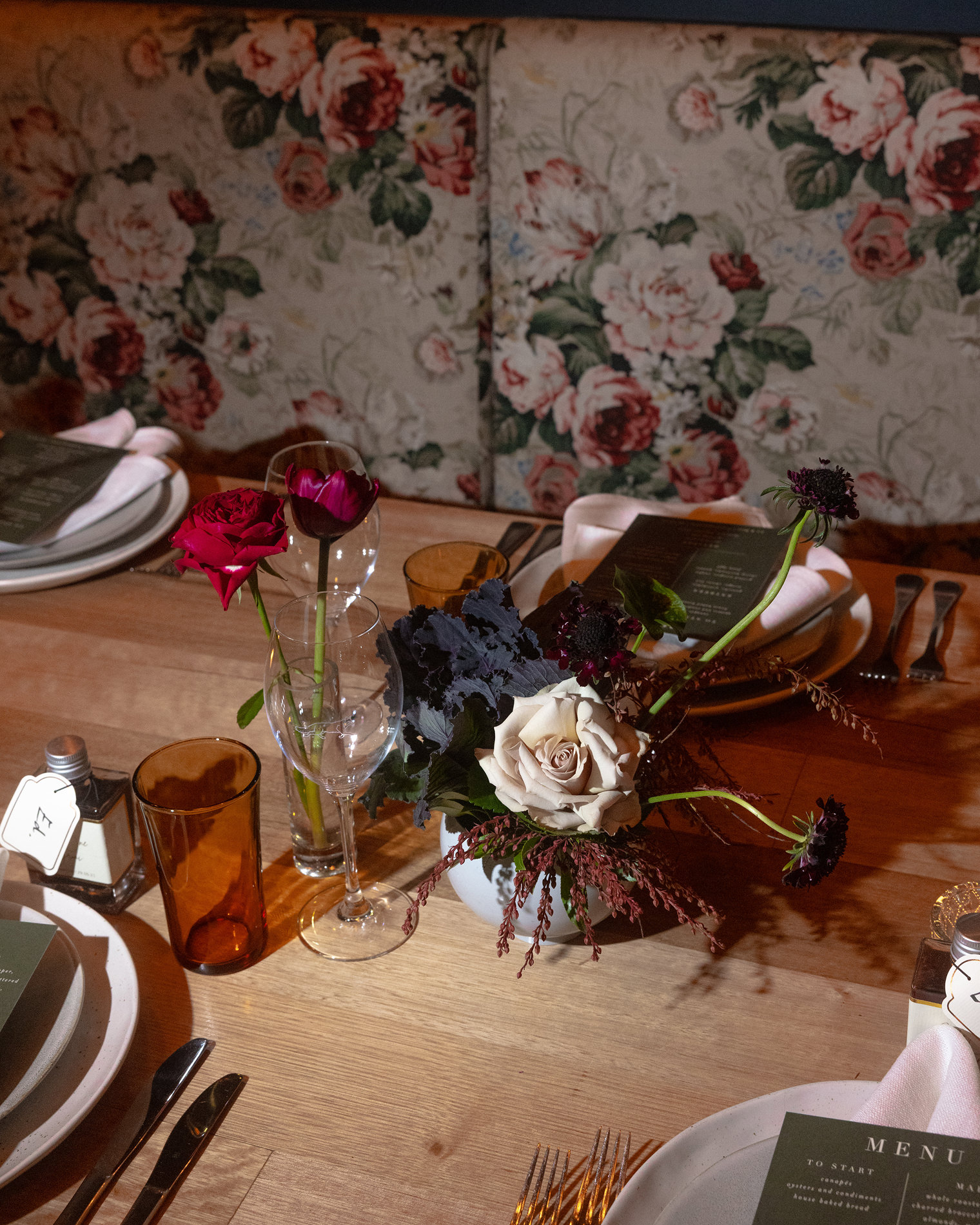 An intimate table setting at a Rupert on Rupert wedding in Melbourne, featuring a rustic wooden table adorned with elegant floral arrangements. Deep red and blush roses, accompanied by dark foliage, are arranged in a centerpiece, complementing the floral-patterned upholstered seating. The place settings include minimalist tableware with a dark green menu card, amber glasses, and neatly folded napkins, creating a warm and inviting atmosphere.