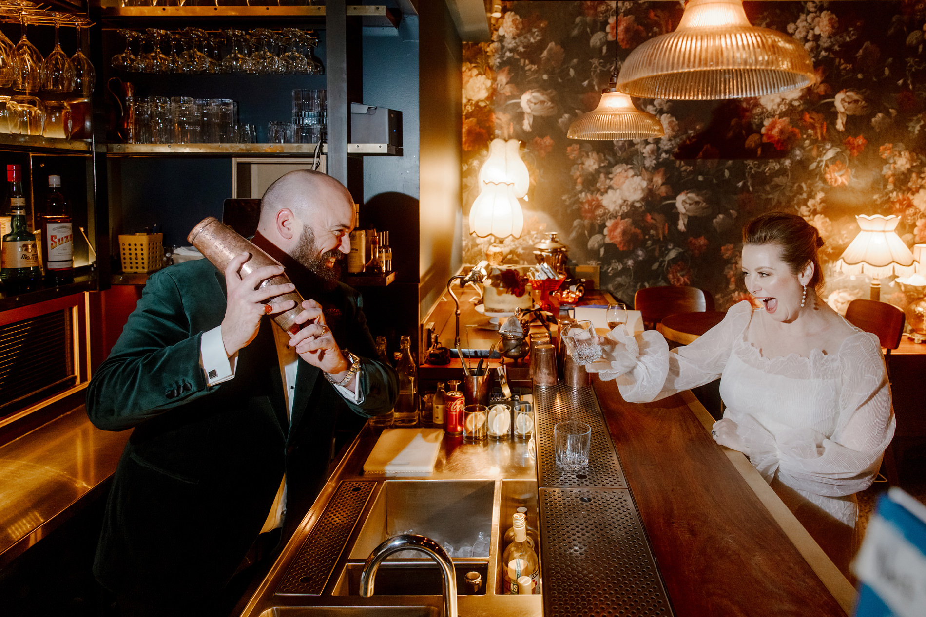 A playful moment at a wedding reception in Rupert on Rupert, Melbourne, captured in a cozy, dimly lit bar with floral wallpaper. The groom, dressed in a velvet jacket, shakes a cocktail shaker with a grin, while the bride, in an elegant white dress, enthusiastically holds up a glass, both sharing a light-hearted laugh.