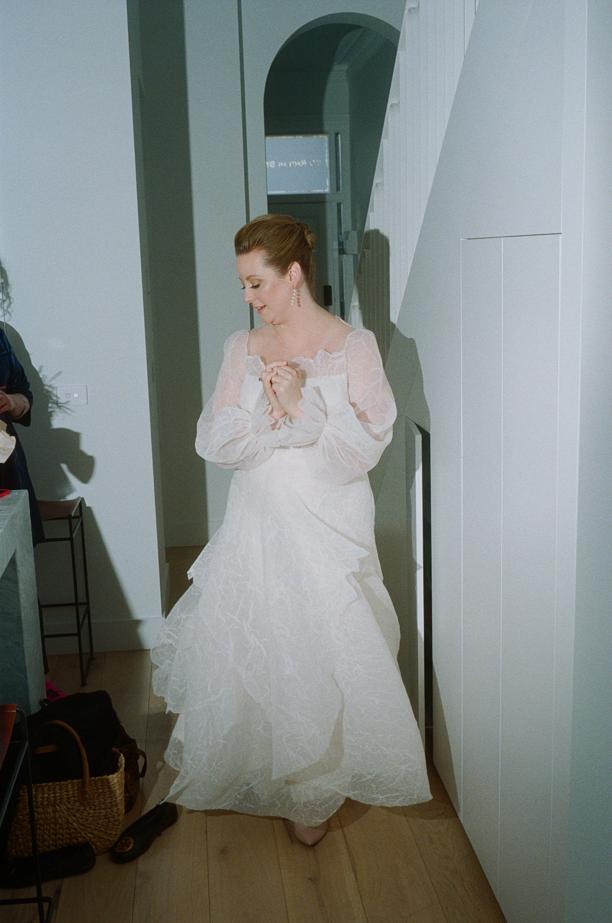 The bride, captured on 35mm Kodak film, is seen standing in a hallway at a Rupert on Rupert wedding in Melbourne. She is wearing a white gown with sheer sleeves and is looking down, clasping her hands together. The scene is set indoors with simple, modern decor visible in the background.