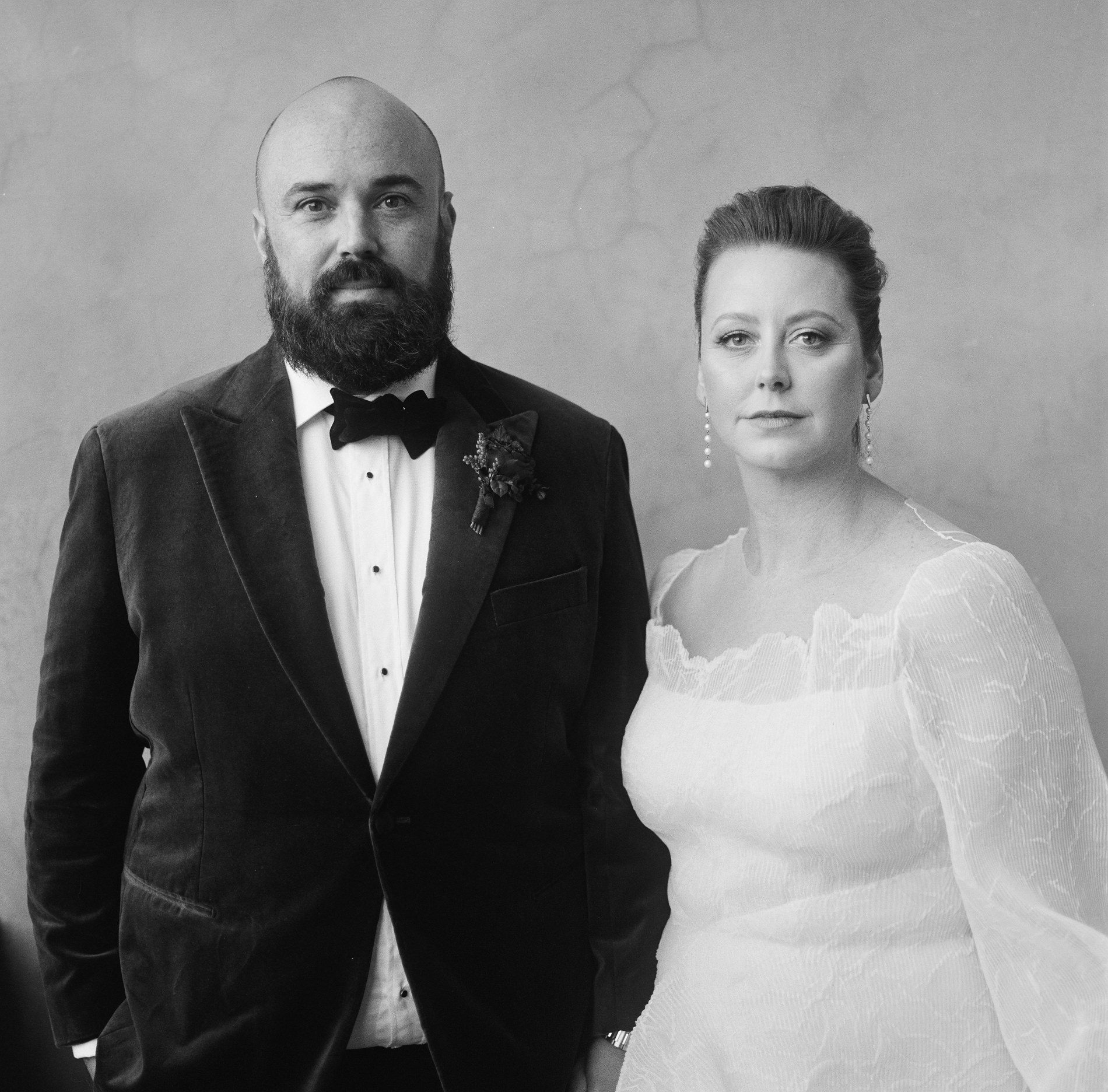 The bride and groom pose for a formal portrait at a Rupert on Rupert wedding in Melbourne, captured on medium-format Kodak film. The groom wears a dark velvet jacket and a bow tie, while the bride, in a white gown with sheer sleeves, stands beside him. Both have neutral expressions as they face the camera against a plain backdrop.