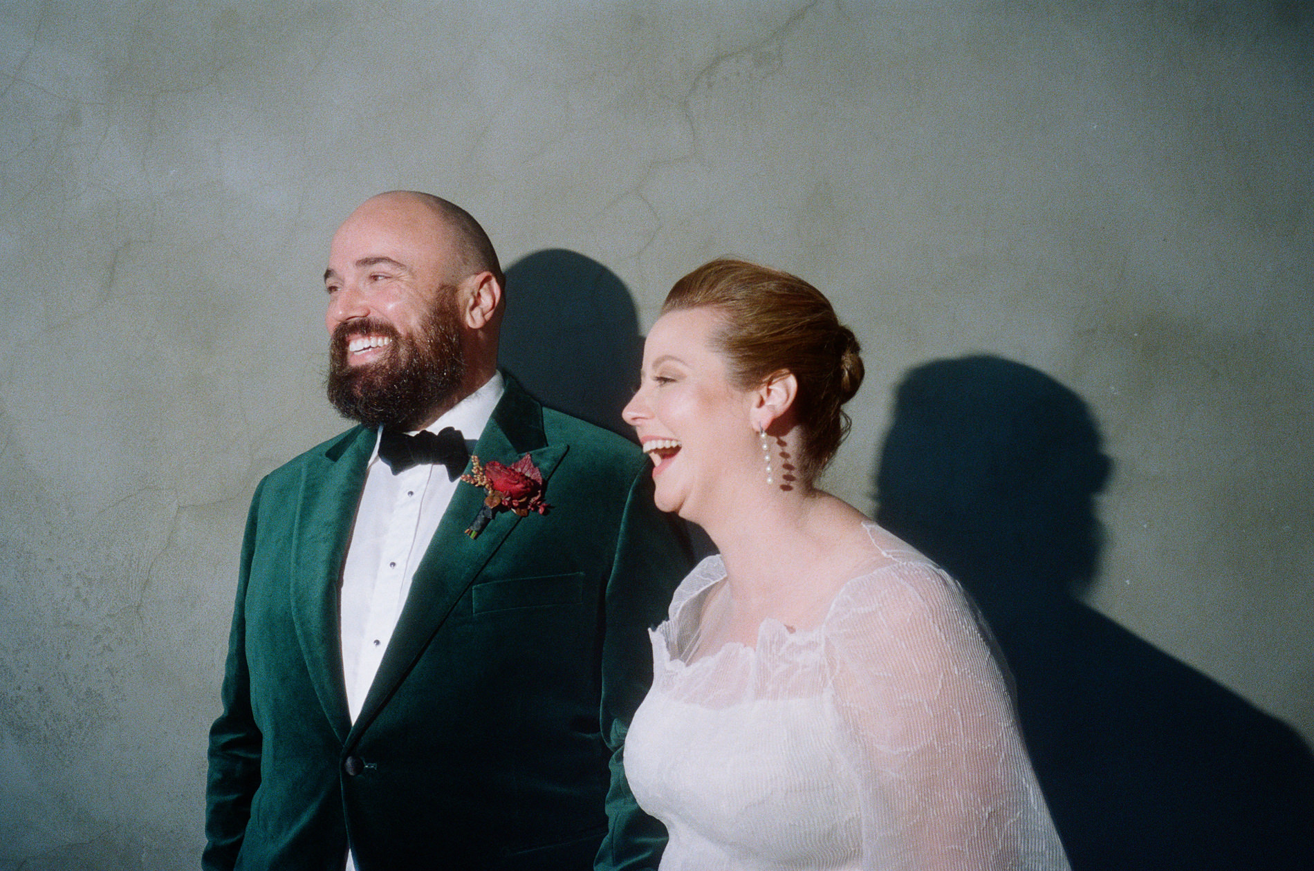 The bride and groom stand against a plain wall at a Rupert on Rupert wedding in Melbourne, captured on 35mm Kodak film. The groom wears a green velvet jacket with a boutonnière, while the bride, dressed in a white gown, smiles broadly. Both are looking off-camera, sharing a lighthearted moment.