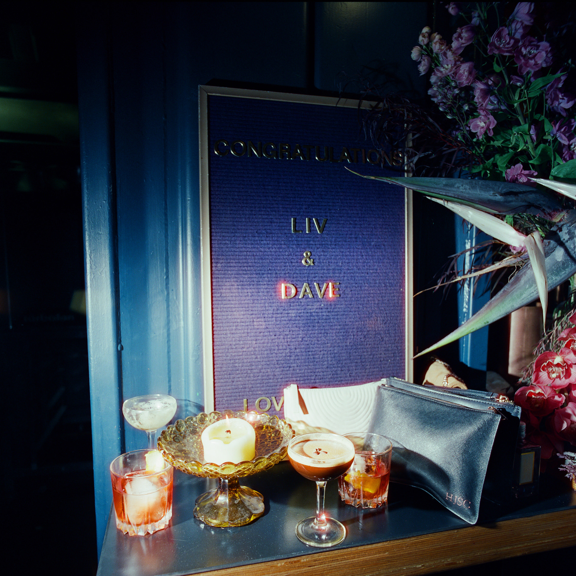 A display at a Rupert on Rupert wedding in Melbourne, featuring a sign that reads 'Congratulations Liv & Dave.' The setup includes several cocktails, a candle in a decorative holder, and a small clutch bag, arranged on a table with flowers in the background.