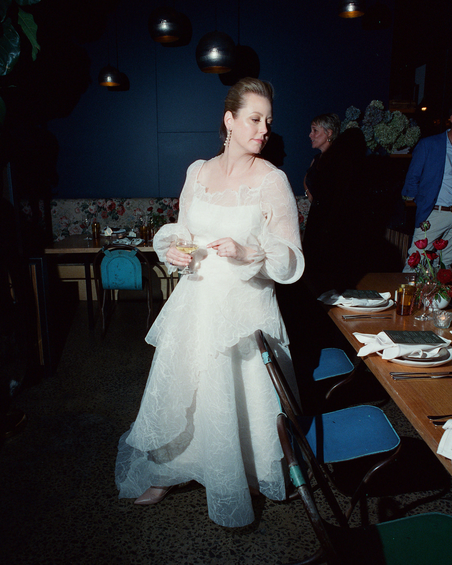 The bride, dressed in a white gown, stands with a drink in hand at a Rupert on Rupert wedding in Melbourne. She is captured in a candid moment, looking off to the side, with tables and chairs partially visible in the background.
