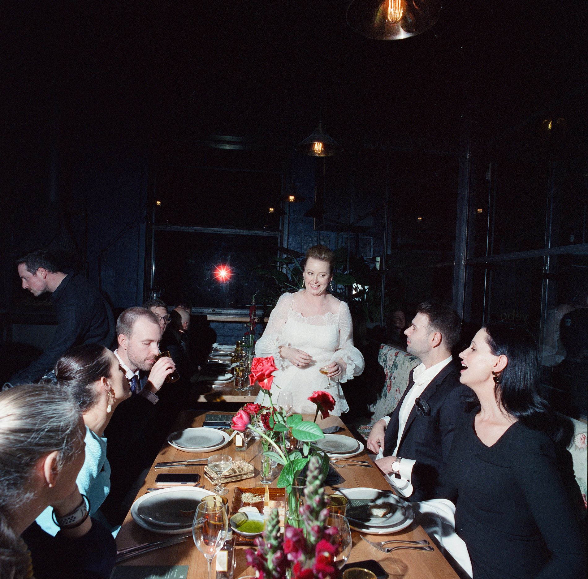 The bride, standing at the head of a long table during a Rupert on Rupert wedding in Melbourne, addresses her seated guests. The table is set for dinner with simple floral arrangements, while guests listen and interact, creating an intimate dining atmosphere.