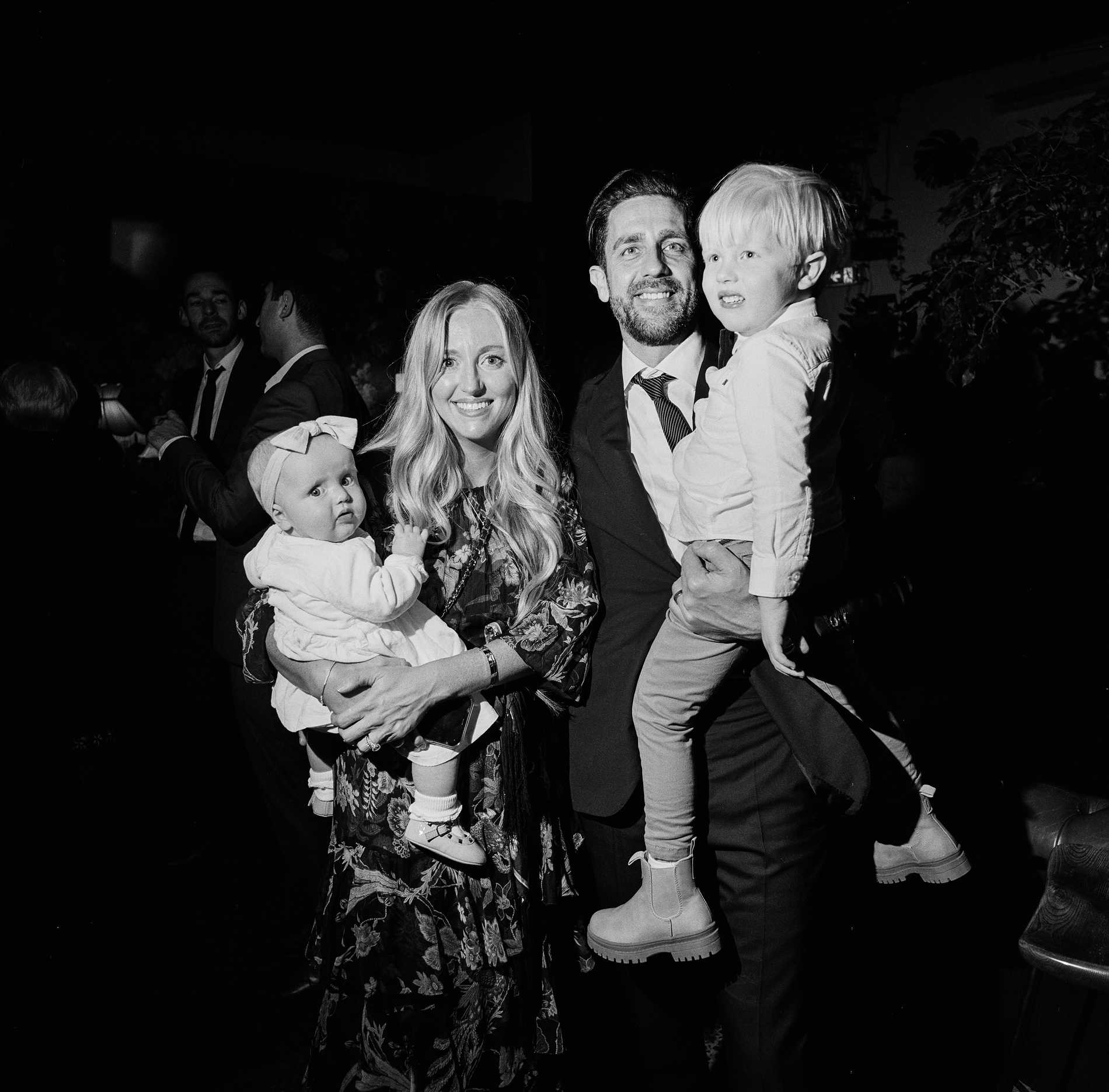 A family portrait taken at a Rupert on Rupert wedding in Melbourne, shot in black and white. A couple poses with their two young children; the father holds the older child while the mother holds the baby. The group is dressed in formal attire, standing against a dark background, with other guests faintly visible behind them.
