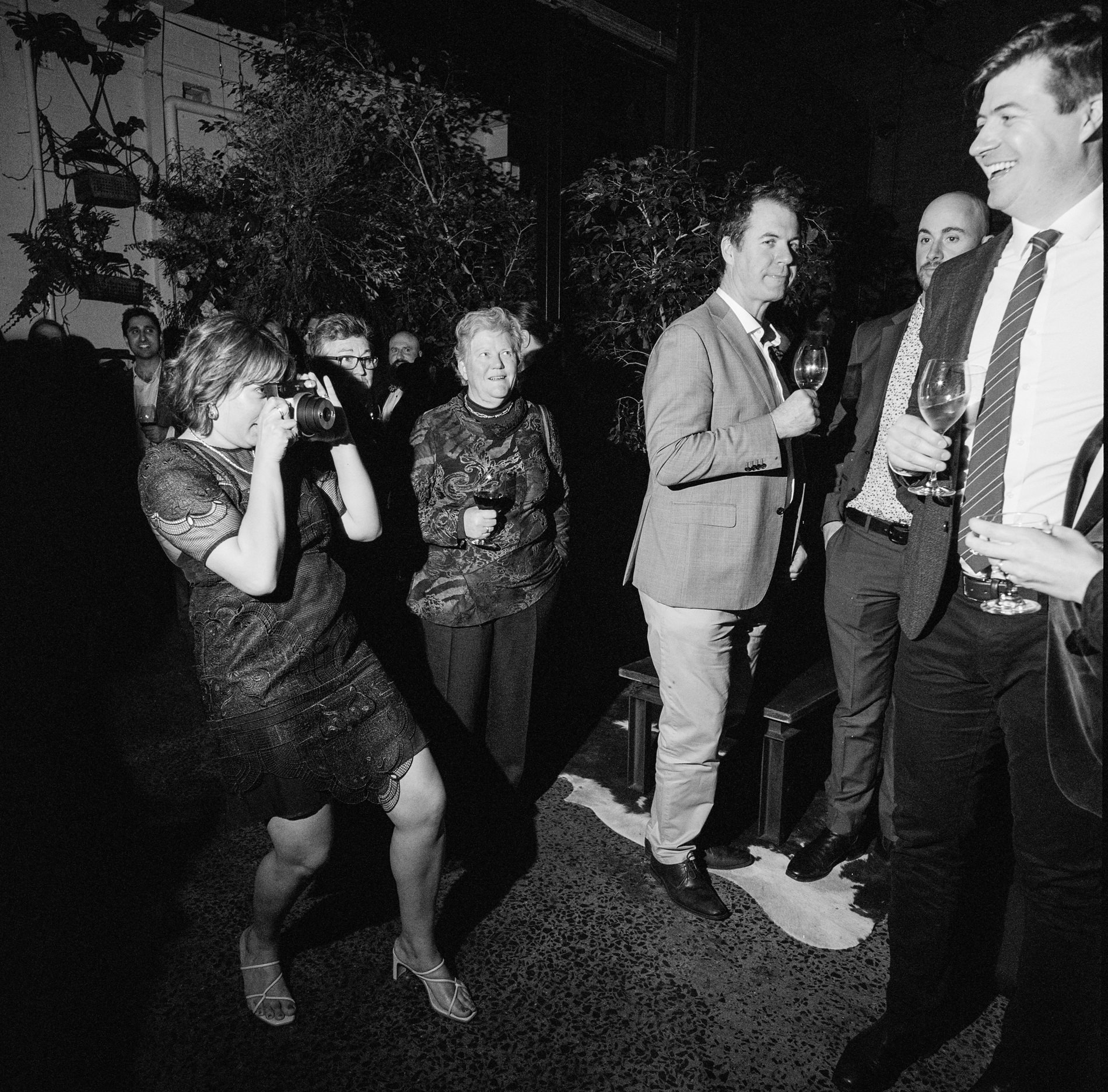 A candid black-and-white moment captured on medium format Kodak film at a Rupert on Rupert wedding in Melbourne. A guest, dressed in a dark lace dress, is seen enthusiastically taking a photo, while other guests in suits and formal attire stand in the background, holding glasses of wine and enjoying the celebration. The scene captures the spontaneous joy and social interaction typical of wedding receptions, with lush greenery in the background adding to the ambiance.