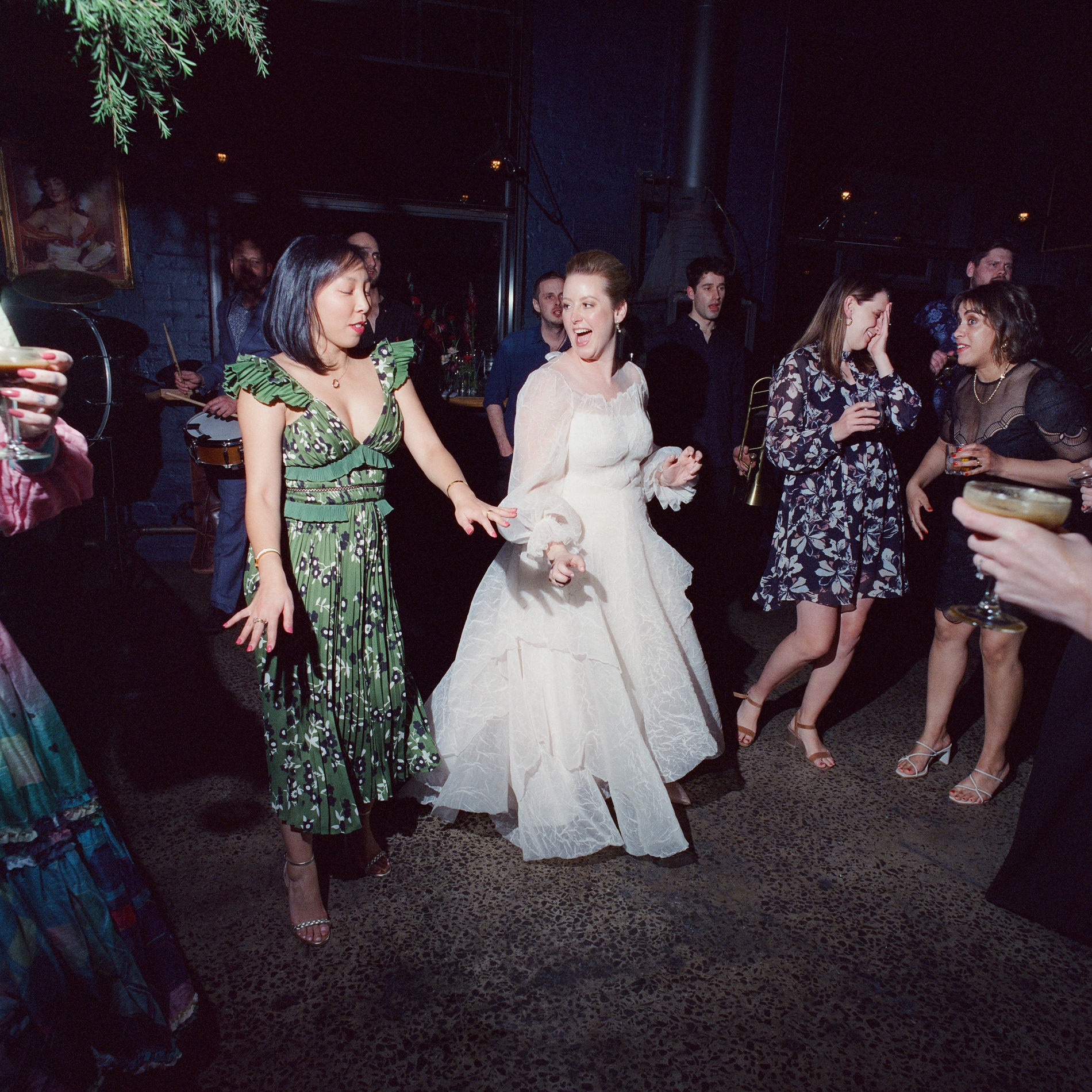 The bride and guests enjoying the dance floor at a Rupert on Rupert wedding in Melbourne. The bride, wearing a flowing white gown with sheer sleeves, dances energetically alongside a guest in a vibrant green dress with ruffled shoulders. The scene captures the lively atmosphere of the celebration, with other guests in the background, drinks in hand, smiling and joining in the fun.