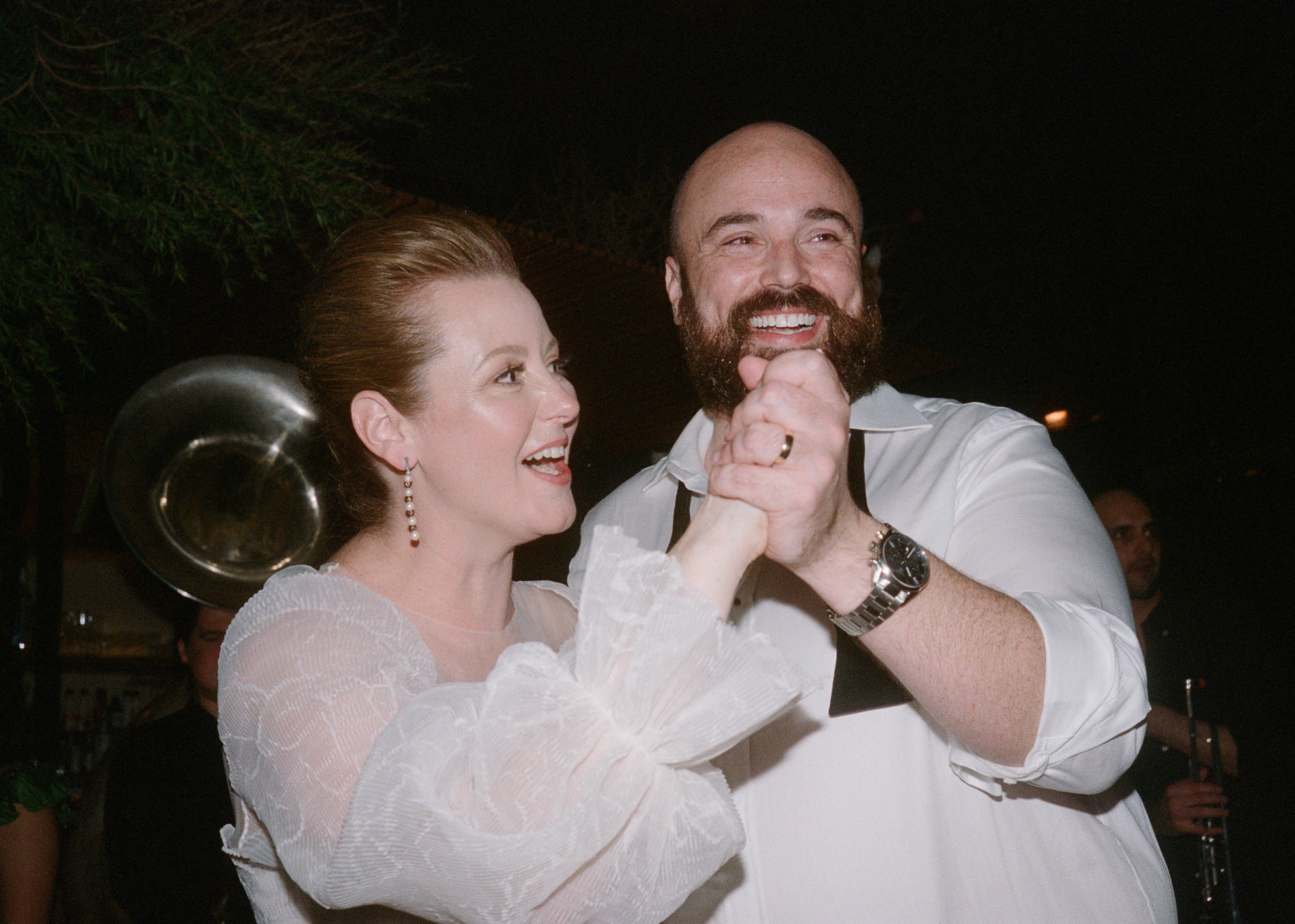A joyous moment captured on 35mm Kodak film at a Rupert on Rupert wedding in Melbourne. The bride and groom are caught mid-dance, laughing and holding hands. The bride, wearing a delicate white dress with sheer sleeves, beams with happiness, while the groom, in a white shirt with his bow tie undone, smiles broadly, embodying the carefree joy of the celebration.