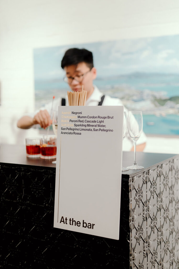 A modern bar setup at a wedding reception in Melbourne, planned by Tori Allen Events. The bar features a stylish black counter with intricate metal detailing, accompanied by a minimalistic drink menu listing options like Negroni, Mumm Cordon Rouge Brut, Peroni Red, and San Pellegrino beverages. In the background, a bartender is seen preparing drinks, adding a dynamic touch to the scene. The setting exudes a sleek and contemporary vibe, with soft lighting and a casual yet refined atmosphere.