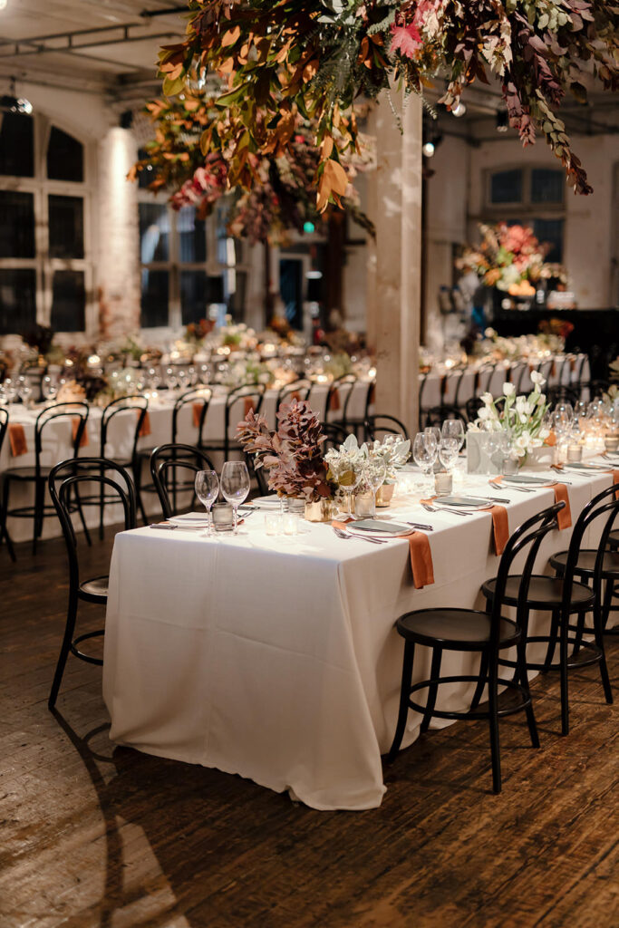 A warmly lit wedding reception venue in Melbourne, designed by Tori Allen Events. The room is adorned with natural autumnal florals and foliage suspended from the ceiling, complementing the soft elegance of long dining tables draped in white linens. The settings are minimalist yet sophisticated, with gold accents, black bentwood chairs, and muted table decor. The overall aesthetic creates an intimate and inviting atmosphere, blending rustic charm with modern elegance.