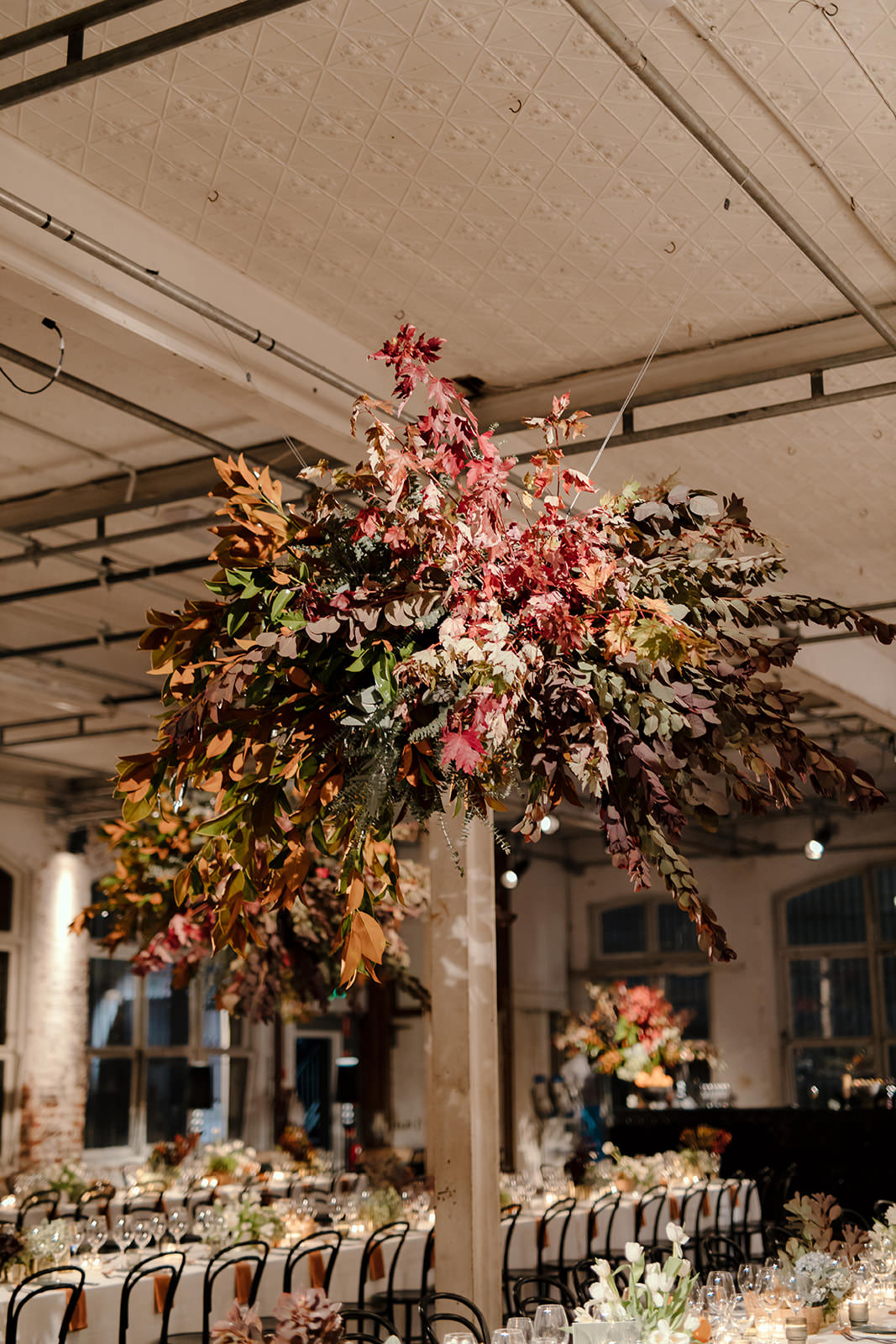 A close-up view of a suspended floral installation at a wedding reception in Melbourne, planned by Tori Allen Events. The arrangement features a striking combination of autumnal foliage in shades of deep red, orange, and green, creating a natural, cascading effect from the ceiling. The room is softly lit, with long tables arranged below, set with elegant glassware, black bentwood chairs, and minimalistic table decor. The ceiling’s industrial framework contrasts with the organic beauty of the floral design, blending modern and rustic aesthetics.