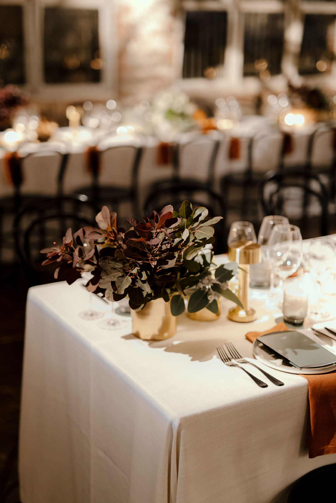 A close-up of a beautifully set table at a wedding reception in Melbourne, planned by Tori Allen Events. The table features a minimalistic arrangement with a small, gold vase holding deep burgundy and green foliage. The soft lighting casts a warm glow on the elegant place settings, which include black plates, silver flatware, and burnt orange napkins that add a pop of color against the white tablecloth. The blurred background showcases additional long tables with matching decor, continuing the understated yet sophisticated aesthetic throughout the venue.