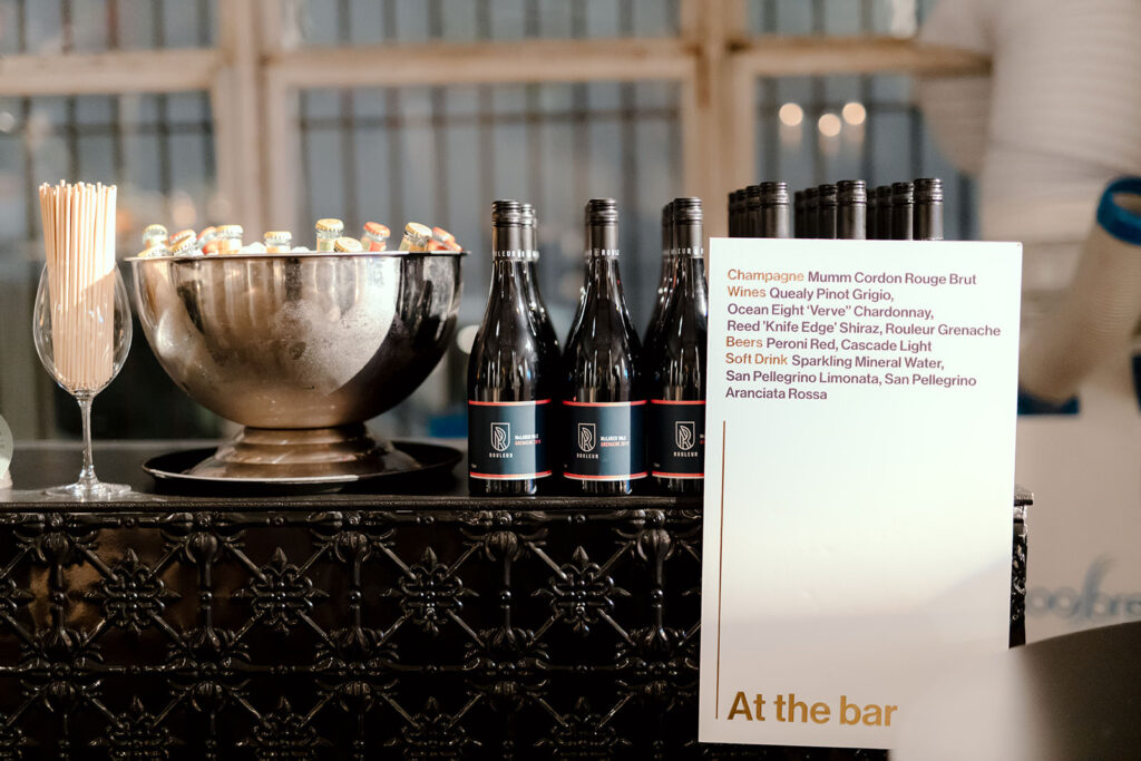 A bar setup at a wedding reception in Melbourne, planned by Tori Allen Events. The display features an elegant drink selection sign listing champagne, wine, beer, and soft drinks available for guests. A metal ice bucket filled with bottled beers sits alongside bottles of wine, with a neatly arranged stack of straws in a wine glass. The sophisticated setup contrasts with the ornate black metal bar front, adding a touch of modern refinement to the venue’s industrial charm.
